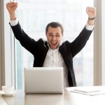 Happy handsome businessman in front of laptop at desk in modern office setting celebrating great news, screaming in joy because of victorious achievement. Successful deal, business success concept.